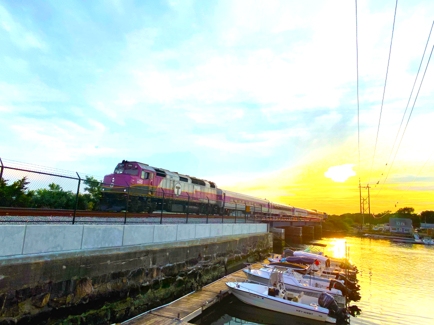 An MBTA Commuter Rail train passes over a bridge at sunset with boats in the foreground
